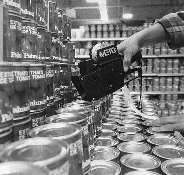 Foto PB de pilhas de latas de estrato de tomate e uma mão segurando uma máquina etiquetadora.