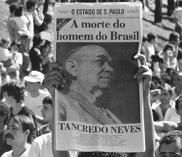 Foto PB. Ao fundo adultos e crianças. Em close, um jornal erguido com a manchete “A morte do homem do Brasil”, a foto de um homem e abaixo, “Tancredo Neves”.