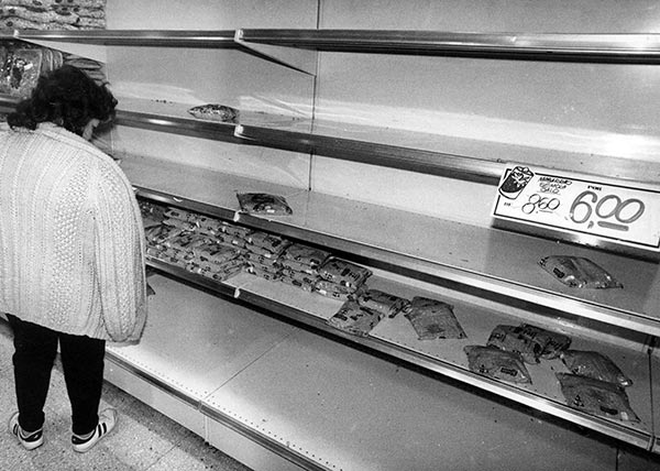 Foto PB. Prateleiras de mercado, à esquerda uma mulher de costas olha para poucos produtos expostos e à direita uma placa “De 8,60 por 6,00”.